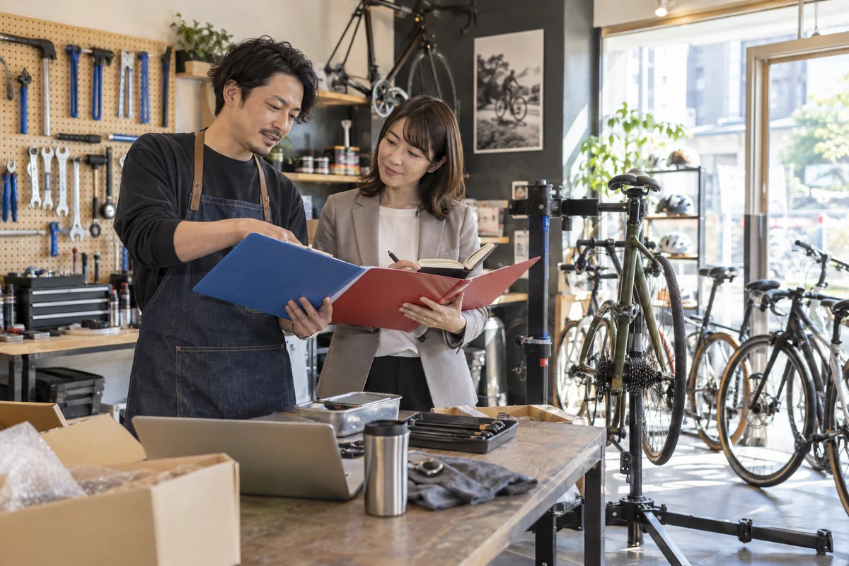 開業準備中の自転車店で設備見積と二つの資金計画を照合する経営者と相談担当者。AI生成の架空イメージ。