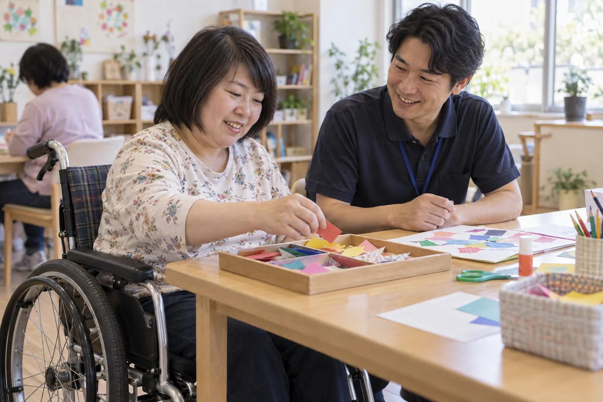 日中の活動室で色紙を選ぶ車椅子の成人と本人の意思を聞く支援員を表したAI生成の架空イメージ