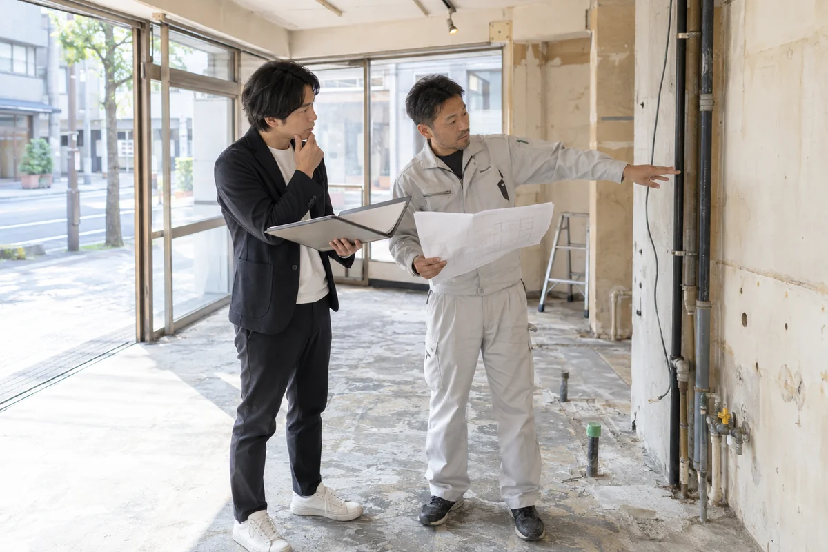 開業予定の空き店舗で創業者と施工担当者が旧物件の精算資料、新物件の図面、工事見積、設備条件を現地確認する架空イメージ